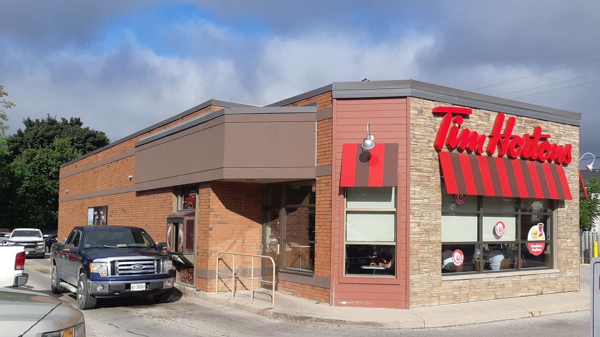 A Tim Hortons storefront in Tottenham, Ontario, with the chain's iconic red signage on the corner of a small brick building and a Ford pickup truck parked at the curb.