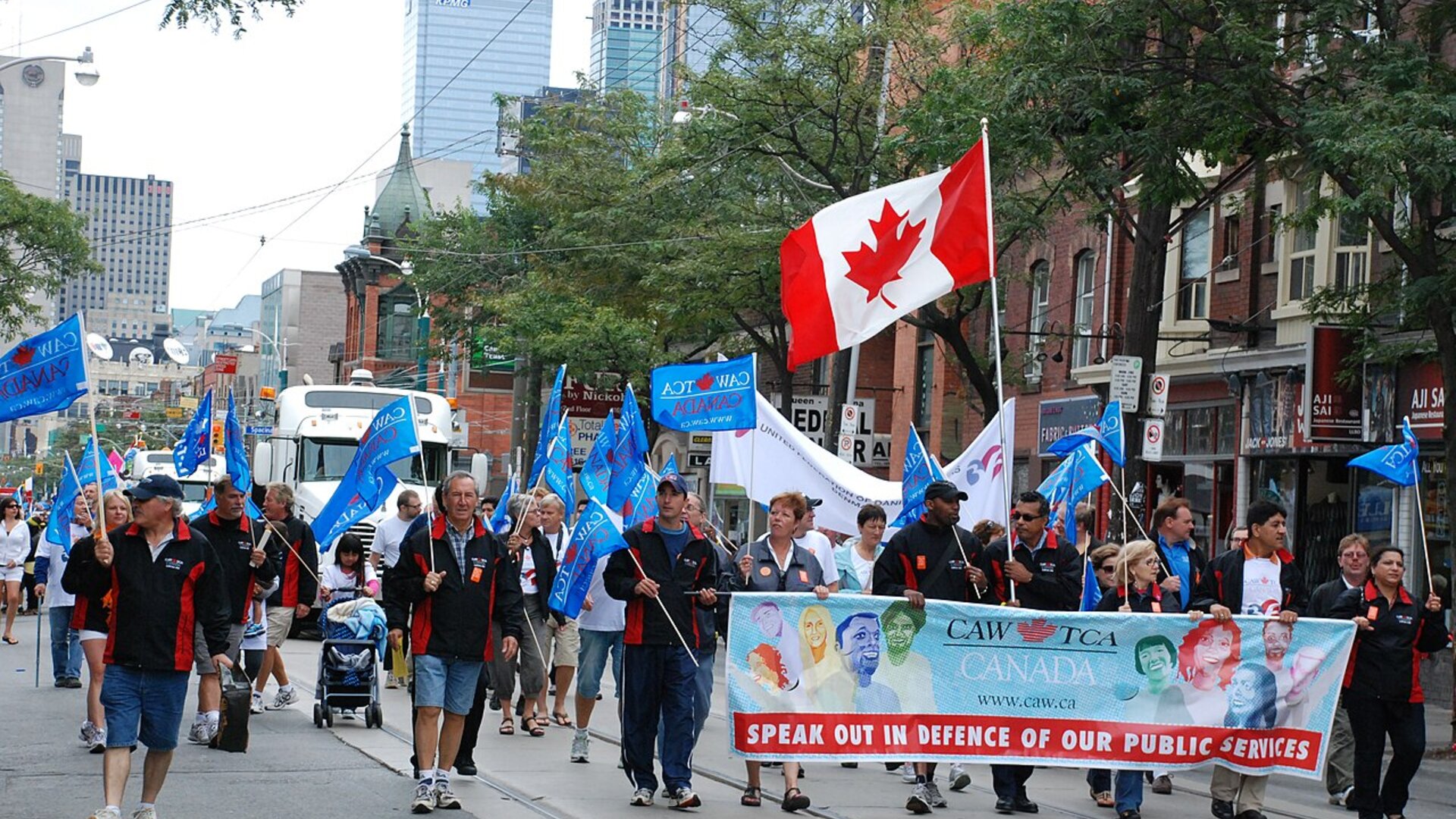 Union members marching in Toronto's 2011 Labour Day Parade behind a banner reading "Speak Out in Defence of Our Public Services," with the downtown skyline behind them.